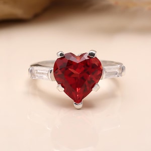 May include: A silver ring with a large, heart-shaped, faceted red gemstone. The ring has two rectangular white gemstones either side of the heart. The ring is set against a soft, neutral background.