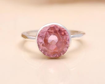May include: A silver ring featuring a large, round, faceted pink gemstone. The gemstone is set in a simple bezel setting, and the band is smooth and polished. The ring is displayed on a light-colored surface.