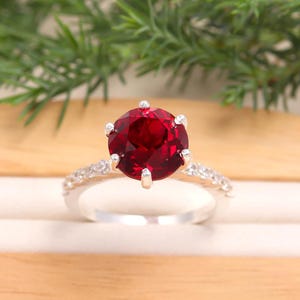 Puede incluir: Un anillo de plata con una gran gema redonda de color rojo intenso como pieza central. La banda está adornada con gemas más pequeñas y transparentes. El anillo se encuentra sobre un fondo neutro con follaje verde.
