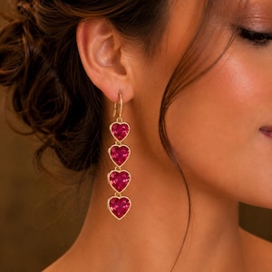 May include: A gold-toned drop earring featuring four vertically aligned, faceted, heart-shaped red stones. Each stone is encased in a gold-colored metal setting, connected by small gold links, hanging from a simple gold-toned hoop ear wire.