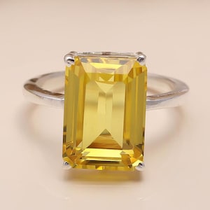 Puede incluir: Un anillo de plata con una gran gema amarilla rectangular. La gema está facetada, captando la luz y mostrando un tono dorado vibrante. La banda del anillo es de plata pulida.
