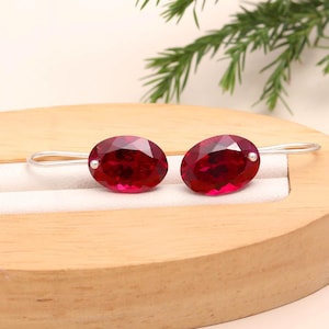 Könnte beinhalten: Ein Paar ovale Ohrringe mit tiefroten Edelsteinen. Die Edelsteine sind in einem silberfarbenen Metall mit einem einfachen Haken-Design gefasst. Die Ohrringe werden auf einer Holzoberfläche präsentiert.