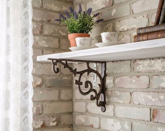 May include: A dark brown metal shelf bracket with an ornate scrollwork design, featuring a curved arm and a vertical mounting plate. The bracket supports a white wooden shelf holding a potted lavender plant, two floral teacups, and books.