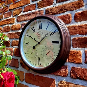 Könnte beinhalten: Eine braune Metall-Außenuhr mit römischen Ziffern und einem Thermometer. Die Uhr ist an einer Backsteinwand montiert.