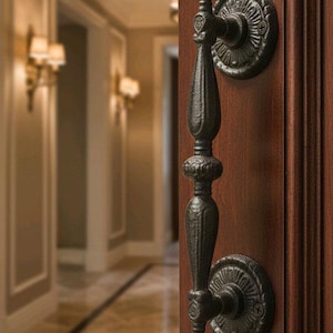 May include: Ornate black door handle with decorative circular backplates, mounted on a rich, dark wood door. The handle has a detailed, textured design, and the background shows a hallway with sconce lighting.