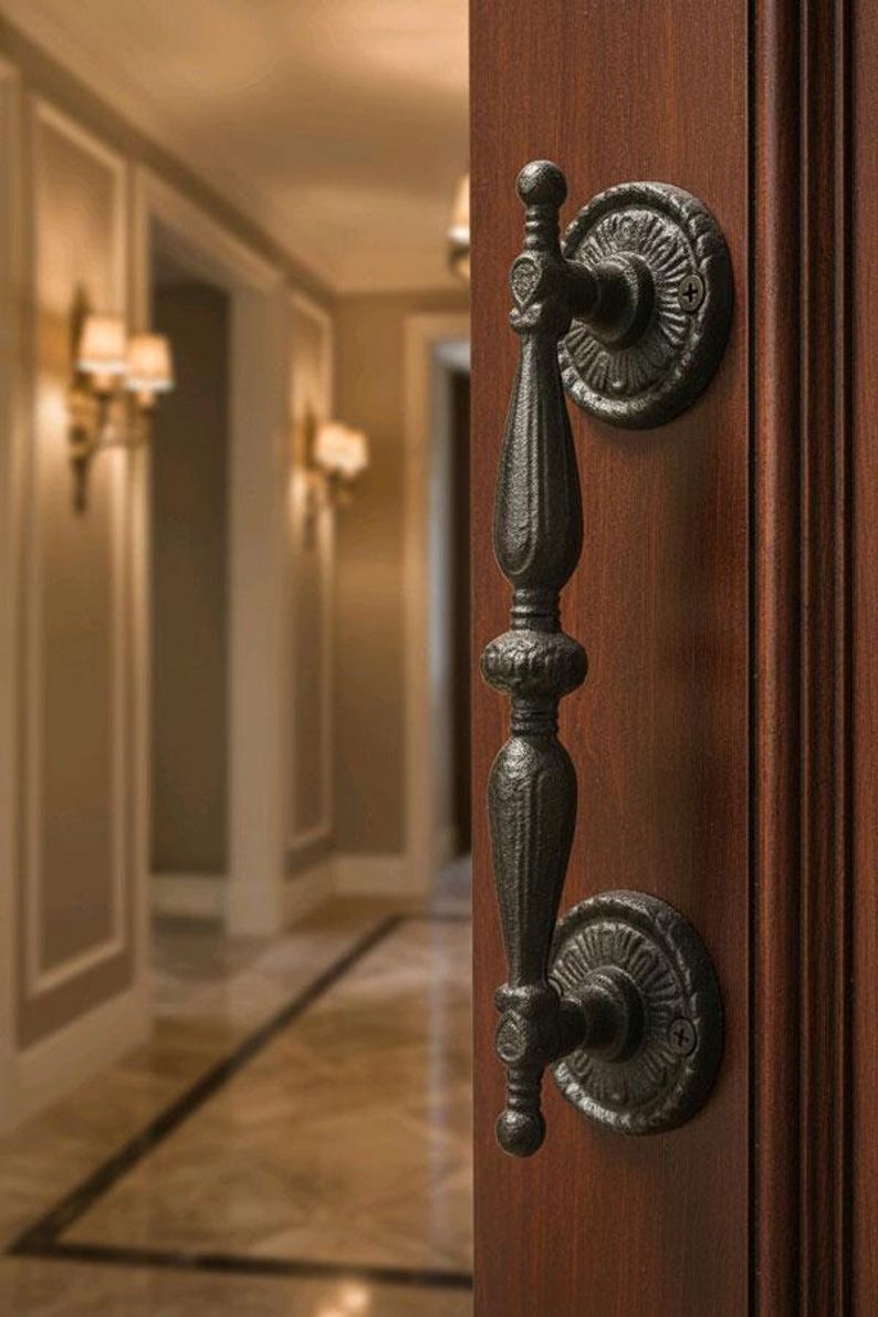May include: Ornate black door handle with decorative circular backplates, mounted on a rich, dark wood door. The handle has a detailed, textured design, and the background shows a hallway with sconce lighting.