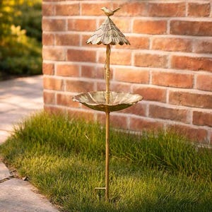 Könnte beinhalten: Ein dekoratives Vogelhaus mit Bronze-Finish. Es hat ein kleines Dach, eine vogelartige Verzierung und eine flache Schale für Futter. Das Vogelhaus steht auf einer schlanken Stange und befindet sich in einem grasbewachsenen Bereich neben einer Backsteinmauer.