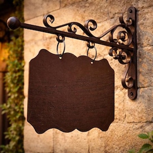 Könnte beinhalten: Ein dunkelbraunes, leeres Schild mit einer dekorativen Halterung aus Schmiedeeisen. Das Schild hat einen gewellten Rand und hängt an einem gebogenen Arm. Die Halterung ist an einer Steinmauer befestigt, mit einer Vintage-Lampe und Grünzeug im Hintergrund.