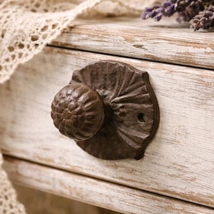 Pode incluir: Close-up de uma gaveta de madeira branca rústica e desgastada com um puxador decorativo de metal marrom escuro. O puxador tem um design redondo e texturizado. Uma renda creme e raminhos de lavanda sugerem uma estética vintage ou rústica.
