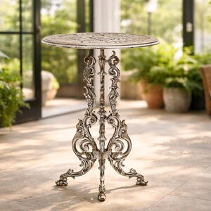 May include: Ornate, antique-style bistro table with a round top and decorative, scrolling legs. The table has a distressed, off-white finish over a metallic base. Ideal for outdoor settings, it measures approximately 28 inches in height.