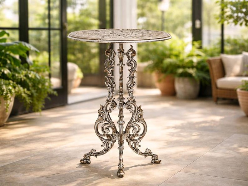 May include: Ornate, antique-style bistro table with a round top and decorative, scrolling legs. The table has a distressed, off-white finish over a metallic base. Ideal for outdoor settings, it measures approximately 28 inches in height.