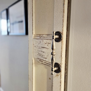 May include: Close-up of a distressed, off-white door with a decorative handle. The door has a textured, aged appearance with visible wear and tear. The handle is ornate, with a dark, circular base and a silver-toned pull.