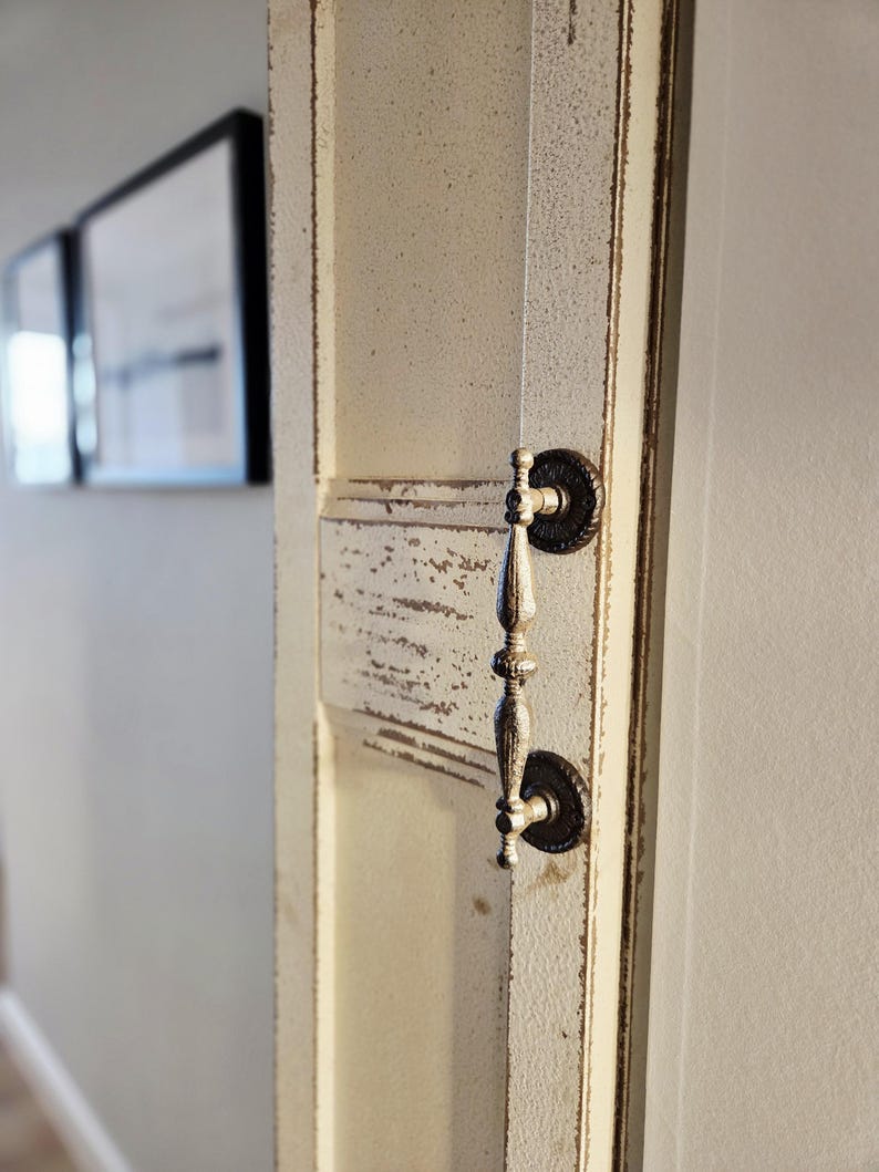 May include: Close-up of a distressed, off-white door with a decorative handle. The door has a textured, aged appearance with visible wear and tear. The handle is ornate, with a dark, circular base and a silver-toned pull.