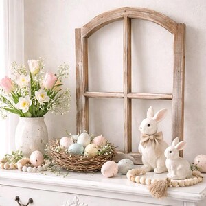 May include: A white dresser is decorated with Easter-themed decor. A bouquet of pink tulips and white daffodils sits in a textured vase. A nest of pastel eggs, two white bunny figurines, and decorative beads complete the display.