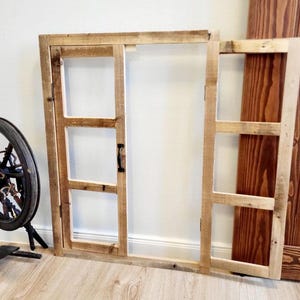 Könnte beinhalten: Ein rustikaler Holzfensterrahmen mit drei Scheiben und einem kleinen Griff. Der Rahmen besteht aus hellem Holz und ist teilweise geöffnet, wodurch eine dunkle Holzplatte sichtbar wird. Im Hintergrund ist ein Spinnrad zu sehen.