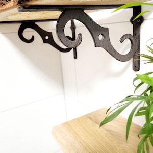 Peut inclure: Étagère en bois rustique avec bord brut, soutenue par deux supports en métal foncé ornés. Les supports présentent un motif en volutes et sont fixés à un mur blanc. Une plante verte est au premier plan.