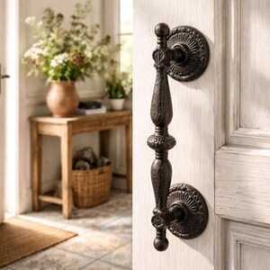May include: Ornate, dark bronze door knocker with a detailed, vertical design. The knocker is mounted on a white-painted wooden door, showcasing a classic, vintage aesthetic. The door is part of a bright, inviting entryway.