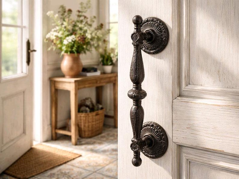 May include: Ornate, dark bronze door knocker with a detailed, vertical design. The knocker is mounted on a white-painted wooden door, showcasing a classic, vintage aesthetic. The door is part of a bright, inviting entryway.