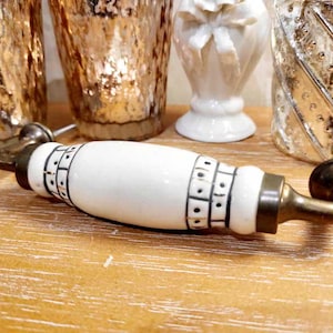 Op de afbeelding: Een witte en messingkleurige kastgreep met een keramisch middenstuk, met een zwart geometrisch patroon. De hardware is ontworpen voor meubels en heeft een vintage esthetiek.