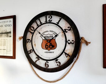 May include: A round, metal wall clock with a rope hanger. The clock face is white with black numbers and a brown and orange Route 66 sign in the center. The clock face has a distressed, vintage look.