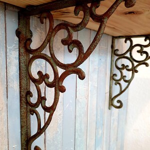 May include: A pair of L-shaped shelf brackets made of cast metal with a weathered, rusted finish and a green patina. The brackets feature an intricate, openwork scroll design with curved metal detailing, supporting a light-coloured wooden shelf.