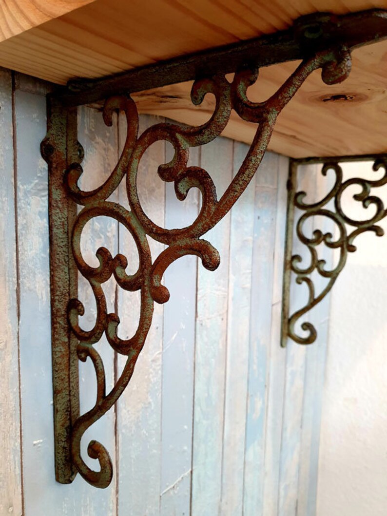 May include: A pair of L-shaped shelf brackets made of cast metal with a weathered, rusted finish and a green patina. The brackets feature an intricate, openwork scroll design with curved metal detailing, supporting a light-coloured wooden shelf.
