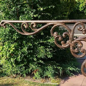 Puede incluir: Soporte de metal ornamental de color bronce con diseños florales y de hojas. El soporte se extiende horizontalmente con un extremo curvado y está montado sobre un soporte vertical. La pieza decorativa se encuentra frente a un follaje verde.