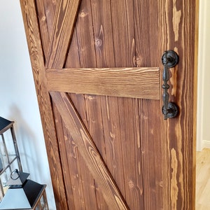 May include: A rustic, wooden barn door with a rich, brown stain. The door features a classic Z-brace design and a black, ornate handle. The wood grain is visible, adding to the door's natural aesthetic. This door is a perfect addition to any home.