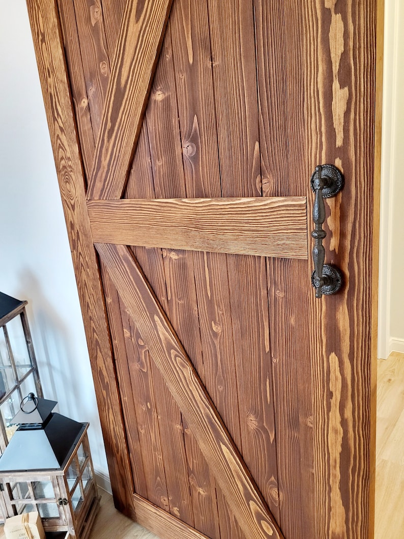 May include: A rustic, wooden barn door with a rich, brown stain. The door features a classic Z-brace design and a black, ornate handle. The wood grain is visible, adding to the door's natural aesthetic. This door is a perfect addition to any home.
