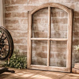 May include: A rustic wooden window frame with an arched top leans against a brick wall. A spinning wheel with a floral arrangement sits to the left. A small vase of flowers and stacked books are on the right. The scene evokes a vintage, farmhouse aesthetic.