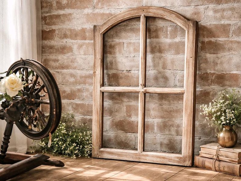 May include: A rustic wooden window frame with an arched top leans against a brick wall. A spinning wheel with a floral arrangement sits to the left. A small vase of flowers and stacked books are on the right. The scene evokes a vintage, farmhouse aesthetic.