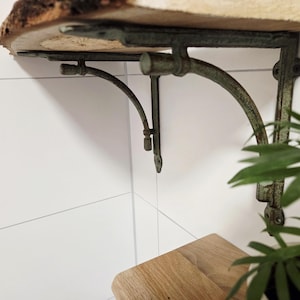 Könnte beinhalten: Rustikales Holzregal mit Baumkante, getragen von zwei geschwungenen Metallhalterungen im Antik-Stil. Die Halterungen sind in einem verwitterten Grünton gehalten und an einer weißen Wand montiert. Eine kleine Pflanze ist in der Ecke sichtbar.