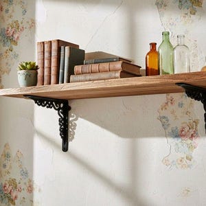 Può includere: Una mensola in legno sostenuta da due staffe in metallo nero decorate. La mensola ospita una piccola pianta succulenta verde in un vaso chiaro, una fila di libri vintage con copertine marroni e grigie e tre bottiglie di vetro colorate.