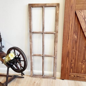 Puede incluir: Una rueca de madera rústica con un arreglo floral color crema. También se ve un marco de ventana rectangular desgastado con seis cristales y una sección de una puerta de madera teñida. La puerta tiene un refuerzo diagonal.