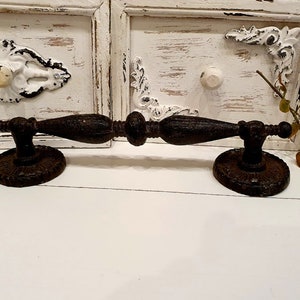 May include: A rustic, dark brown metal drawer pull with an ornate design. The pull features a central handle with decorative detailing and is mounted on two circular bases. The background includes white painted cabinet doors with distressed accents.