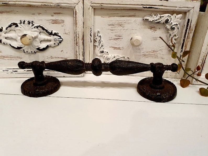 May include: A rustic, dark brown metal drawer pull with an ornate design. The pull features a central handle with decorative detailing and is mounted on two circular bases. The background includes white painted cabinet doors with distressed accents.