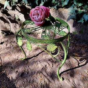 Pode incluir: Um pequeno e ornamentado suporte de plantas de metal verde com um tampo redondo e pernas curvas. O suporte apresenta detalhes decorativos de folhas e volutas. Uma rosa rosa repousa sobre o suporte. Ideal para exibir pequenas plantas ou objetos decorativos. Mede cerca de 30 cm de diâmetro.
