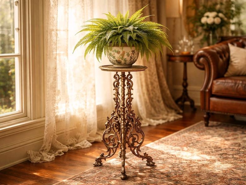 May include: A decorative plant stand with a fern in a floral patterned pot. The stand is made of ornate, bronze-colored metal and has a round top. The fern has long, green fronds. The stand is 30 inches tall.