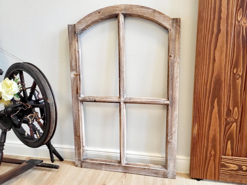 May include: A rustic, arched window frame with a weathered, whitewashed finish. The frame is divided into four panes. A spinning wheel with a floral arrangement is in the background. The window frame is made of wood.