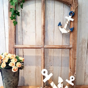 May include: A rustic wooden window frame with a decorative nautical theme. Two white anchors with rope detailing and blue accents are displayed. A vase of orange roses and green ivy add to the coastal aesthetic.