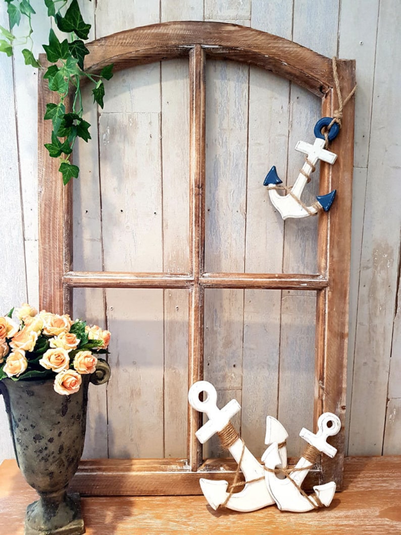 May include: A rustic wooden window frame with a decorative nautical theme. Two white anchors with rope detailing and blue accents are displayed. A vase of orange roses and green ivy add to the coastal aesthetic.