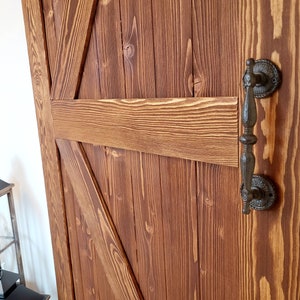 May include: Close-up of a rustic wooden barn door with a dark brown finish. The door features a decorative black metal handle and a classic cross-brace design. The wood grain is visible, adding to the door's charm.