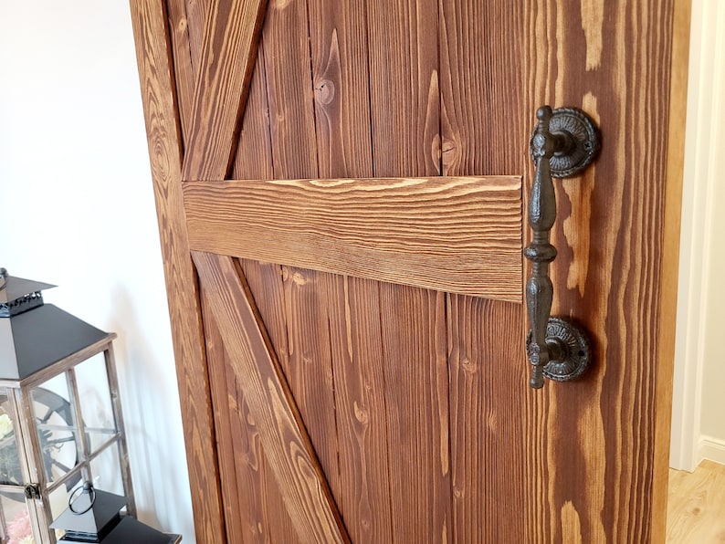 May include: Close-up of a rustic wooden barn door with a dark brown finish. The door features a decorative black metal handle and a classic cross-brace design. The wood grain is visible, adding to the door's charm.