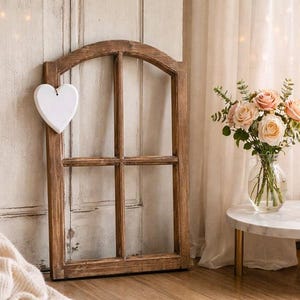 May include: A rustic wooden window frame with a white heart attached. A bouquet of pink and white roses sits in a glass vase on a small marble table with gold legs. Two lit candles in gold holders add warmth to the scene.
