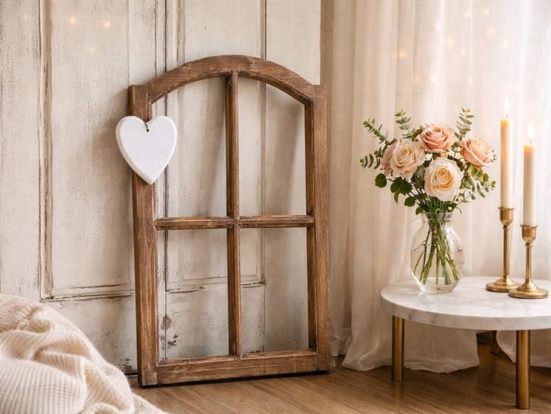 May include: A rustic wooden window frame with a white heart attached. A bouquet of pink and white roses sits in a glass vase on a small marble table with gold legs. Two lit candles in gold holders add warmth to the scene.