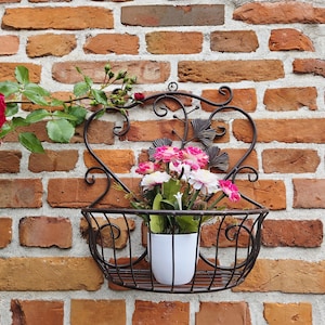 Puede incluir: Una jardinera de pared decorativa de hierro forjado con una parte superior en forma de corazón y una cesta para flores. La jardinera contiene una maceta blanca con flores rosas y blancas contra una pared de ladrillos. Rosas rojas visibles a la izquierda.