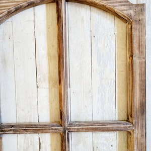May include: A rustic wooden arched window frame with a distressed finish. The frame is constructed from dark brown wood, with a light-colored, vertical plank background. The design features a curved top and a grid pattern.