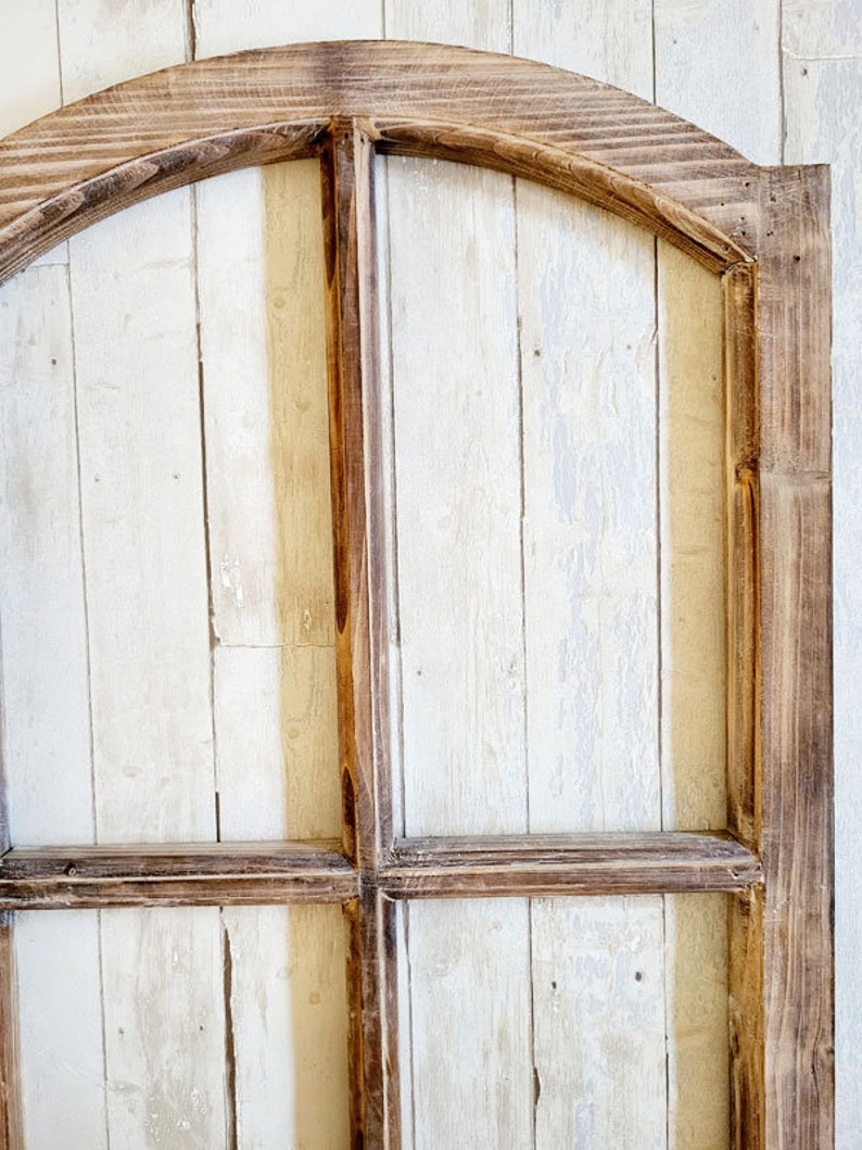 May include: A rustic wooden arched window frame with a distressed finish. The frame is constructed from dark brown wood, with a light-colored, vertical plank background. The design features a curved top and a grid pattern.
