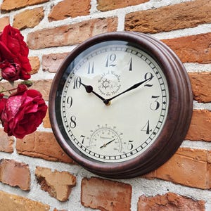 Könnte beinhalten: Eine runde, braun gerahmte Outdoor-Uhr mit cremefarbenem Zifferblatt, an einer Backsteinmauer befestigt. Die Uhr zeigt die Zeit, Temperatur und den Text "World of Weather Temps du Monde". Rote Rosen sind im Vordergrund zu sehen. Der Durchmesser beträgt ca. 25 cm.