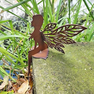Può includere: Una scultura di fata in metallo arrugginito con intricate ali a forma di foglie, seduta su una pietra muschiosa. La fata è una silhouette marrone, su uno sfondo di verde e acqua.